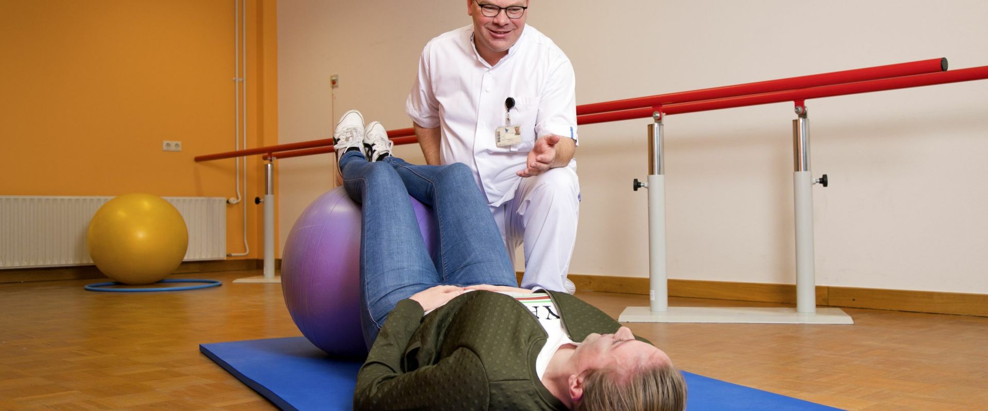 Vrouw doet oefeningenen op een mat met een bal onder begeleiding van een fysiotherapeut.