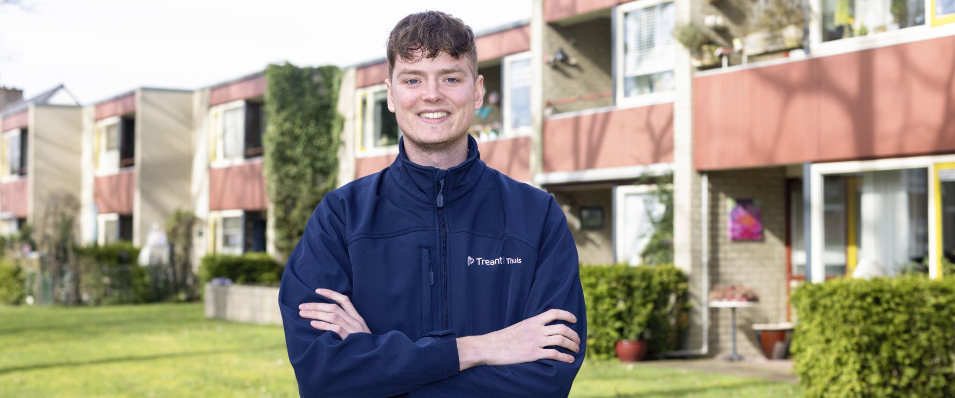 Een persoon staat buiten voor een woonzorglocatie, glimlachend in werkkleding van Treant Thuis