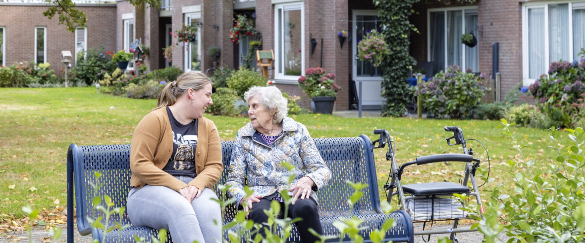 Twee vrouwen zitten op een bankje in een tuin voor een wooncomplex, met een rollator naast hen.