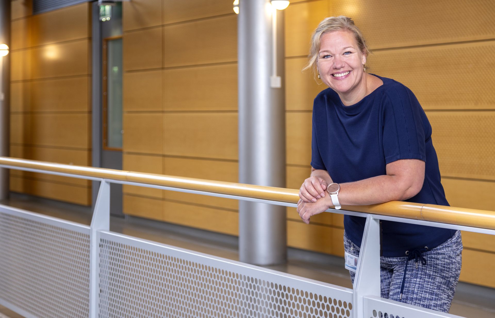 Vrouw leunt glimlachend op een railing in een moderne gang.