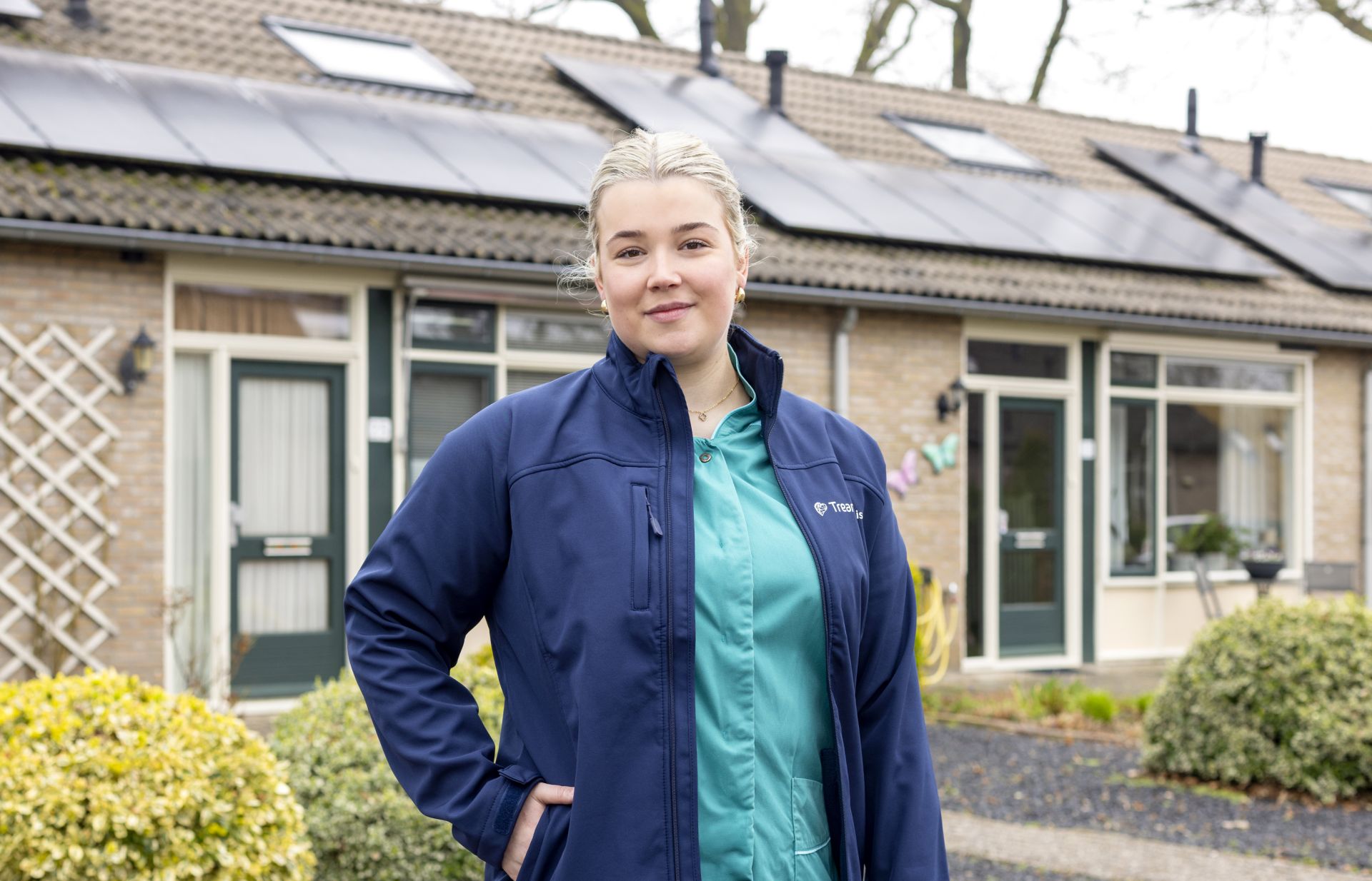 Een vrouw poseert voor een huis in werkkleding van Treant Thuis.