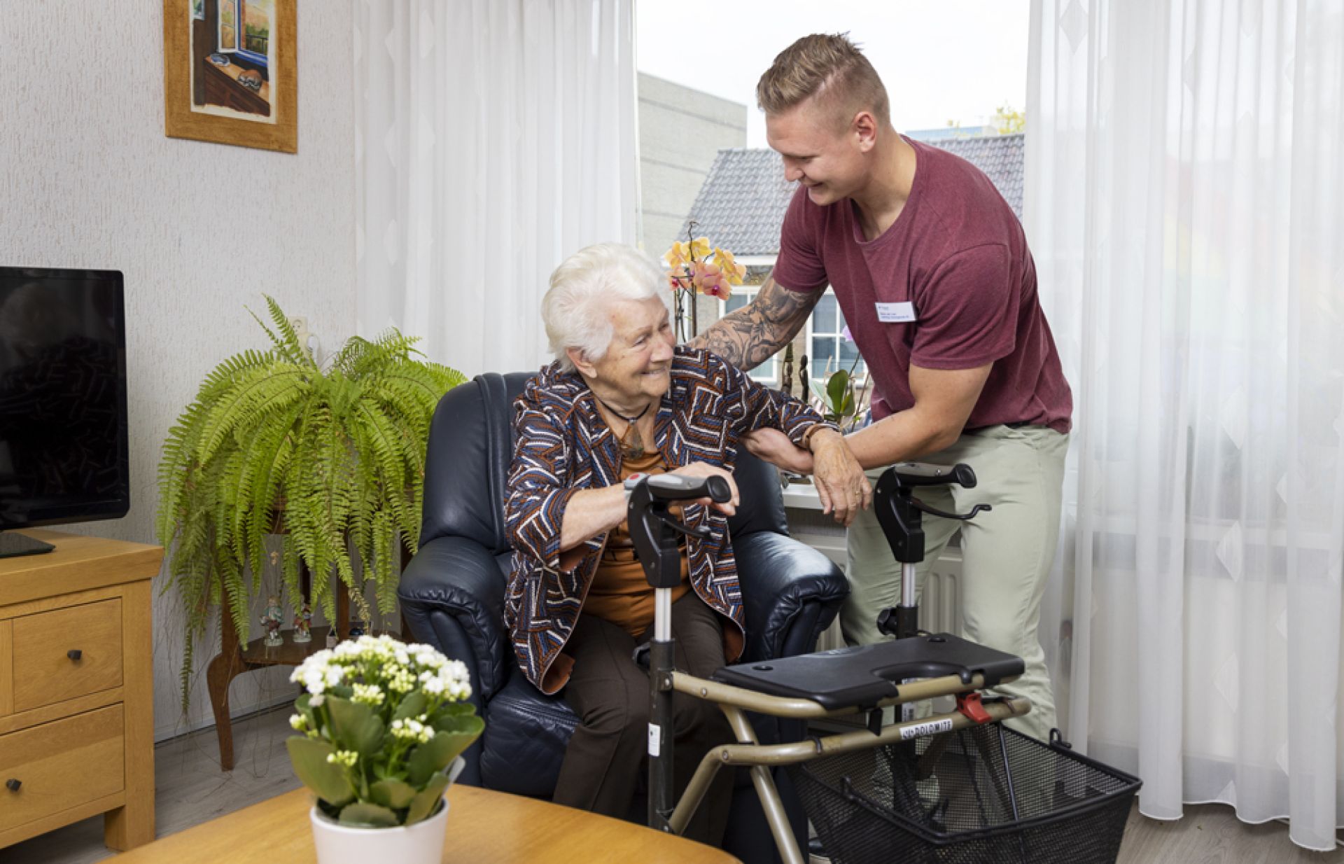 Bewoner krijgt hulp van een medewerker bij het opstaan uit haar stoel.