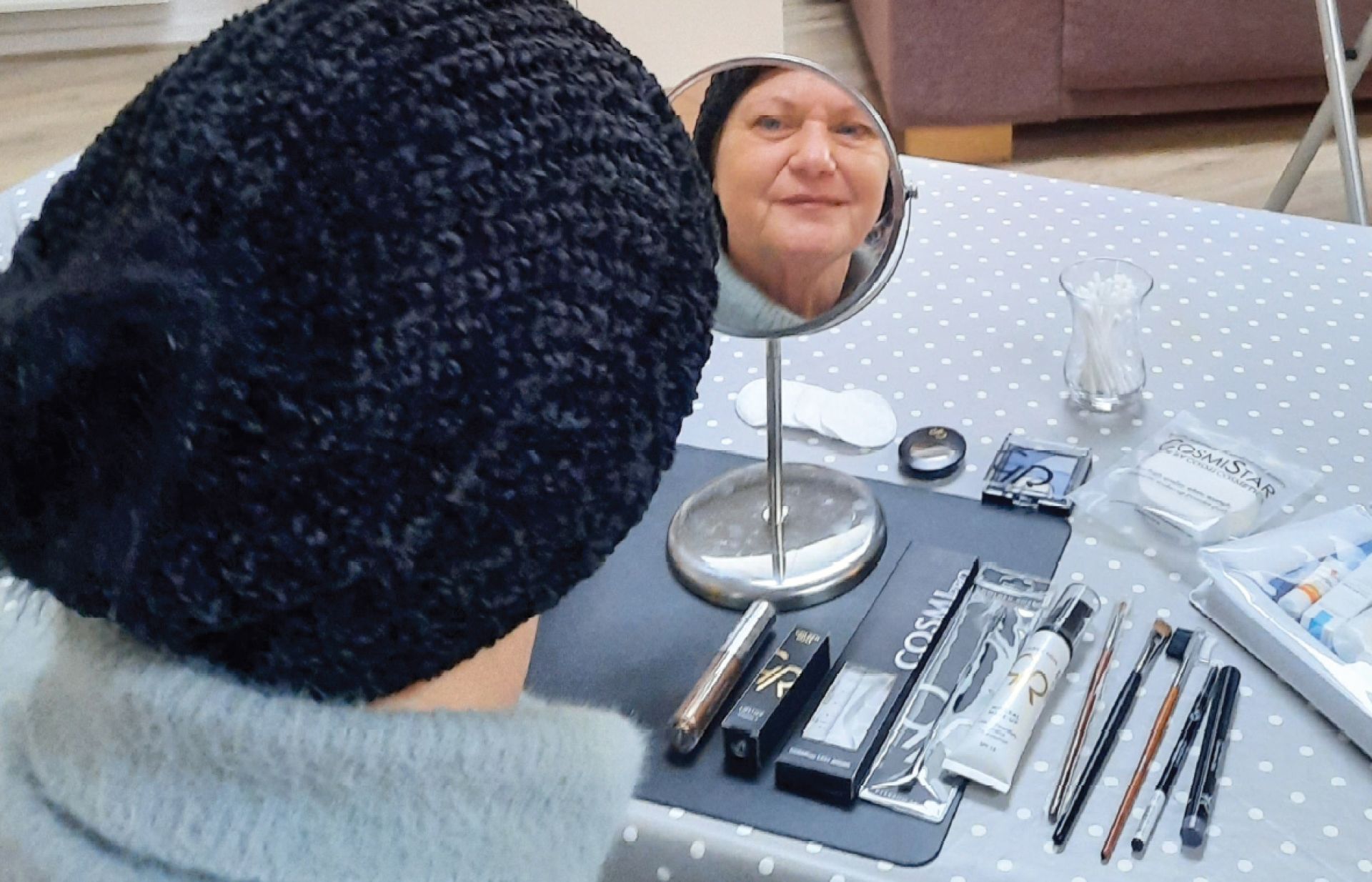 Een mevrouw kijkt in de spiegel. Daaromheen ligt make up. Ze draagt een muts. Je ziet haar reflectie