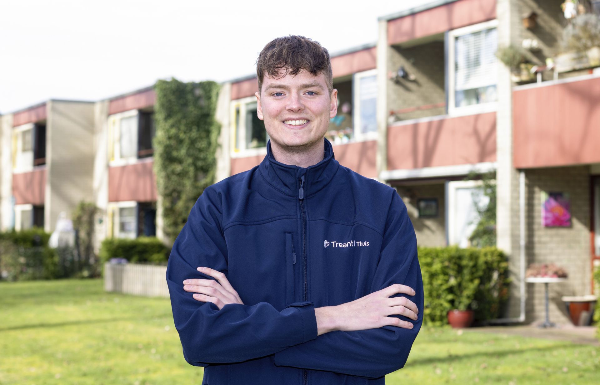 Een persoon staat buiten voor een woonzorglocatie, glimlachend in werkkleding van Treant Thuis
