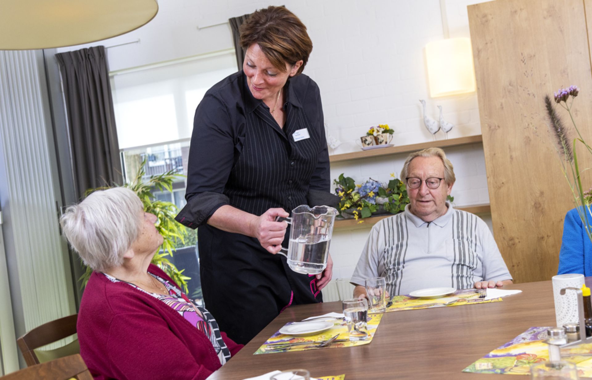 Medewerker voorziet bewoners van een glas water aan de eettafel.