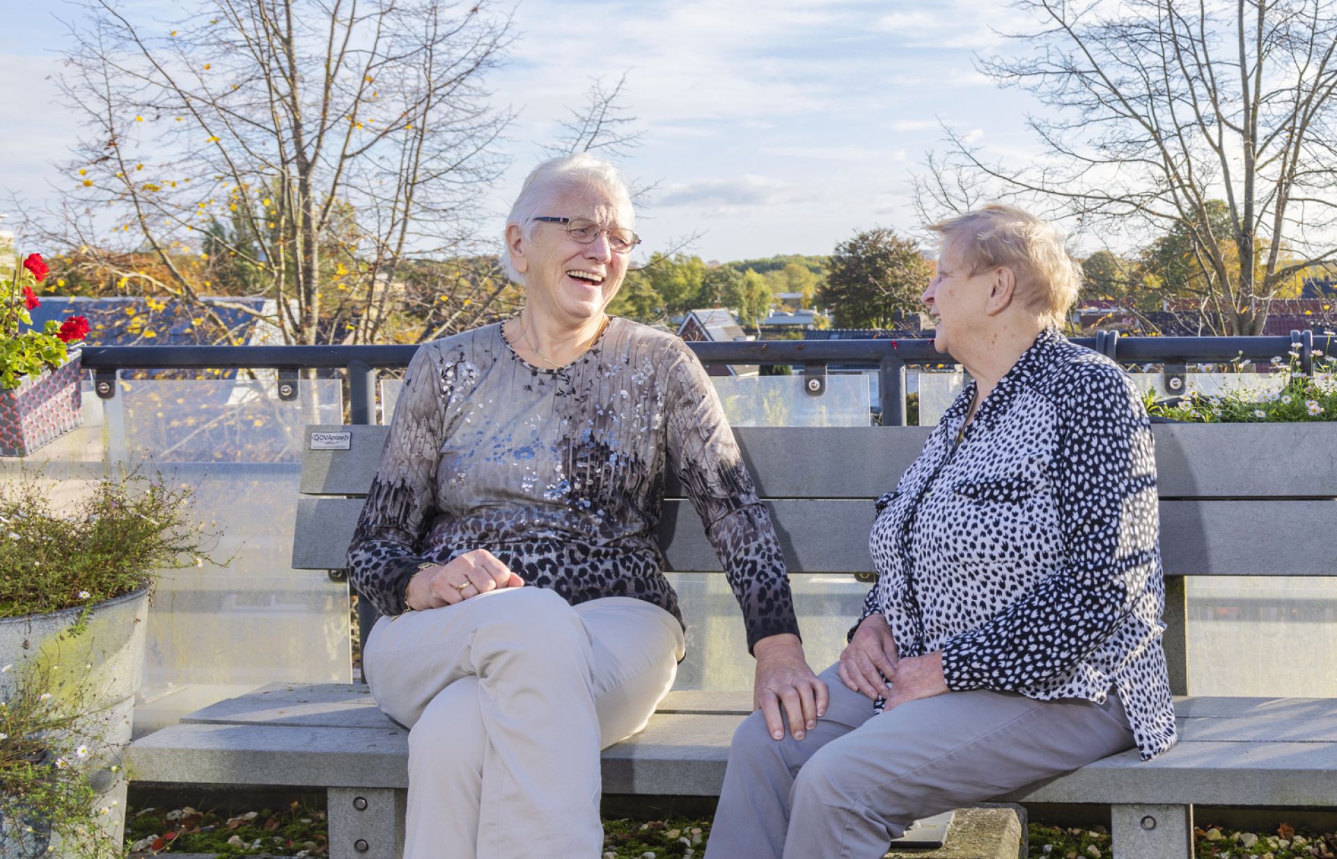 Twee bewoners zitten lachend buiten op een bankje in de zon