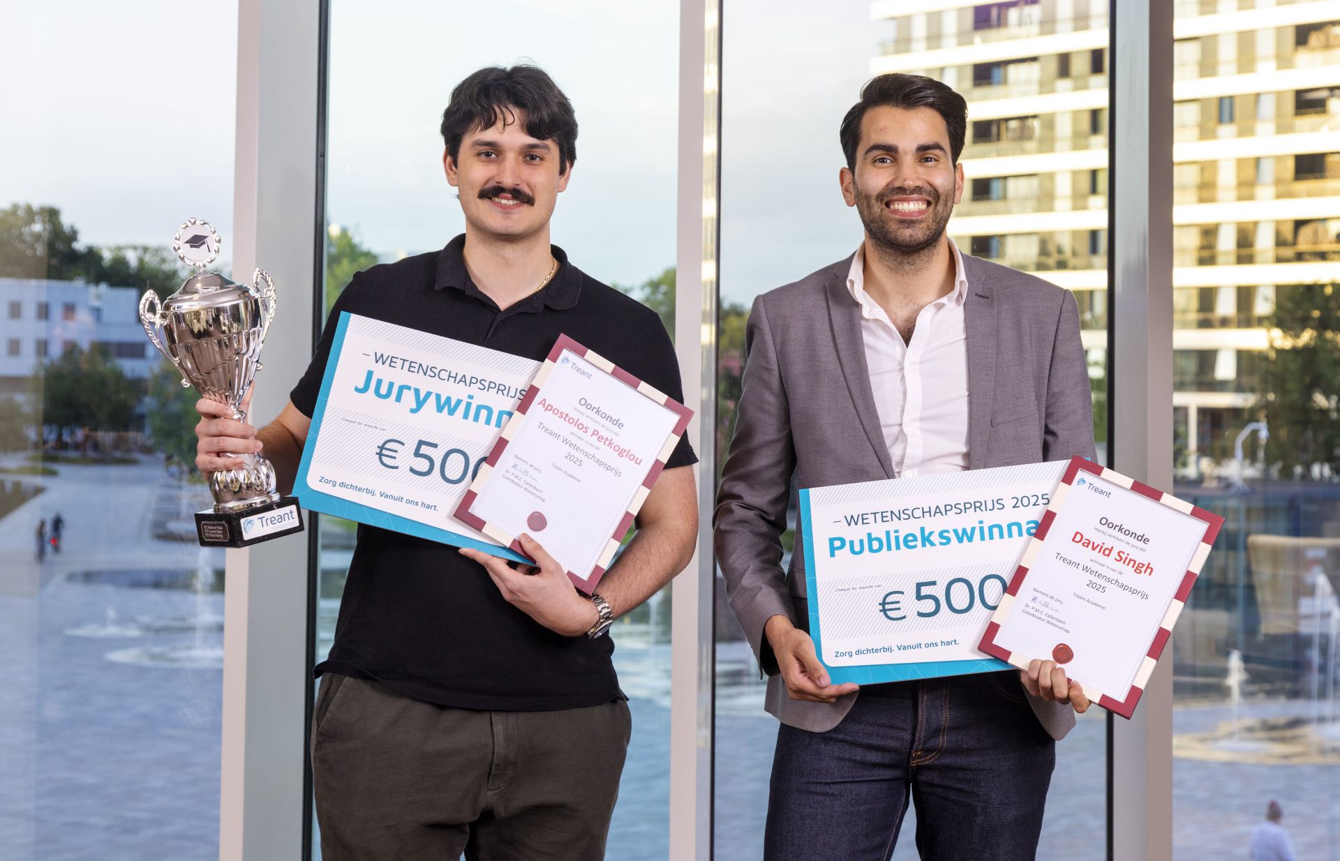 Twee mannen poseren met trofeeën en certificaten voor een raam.