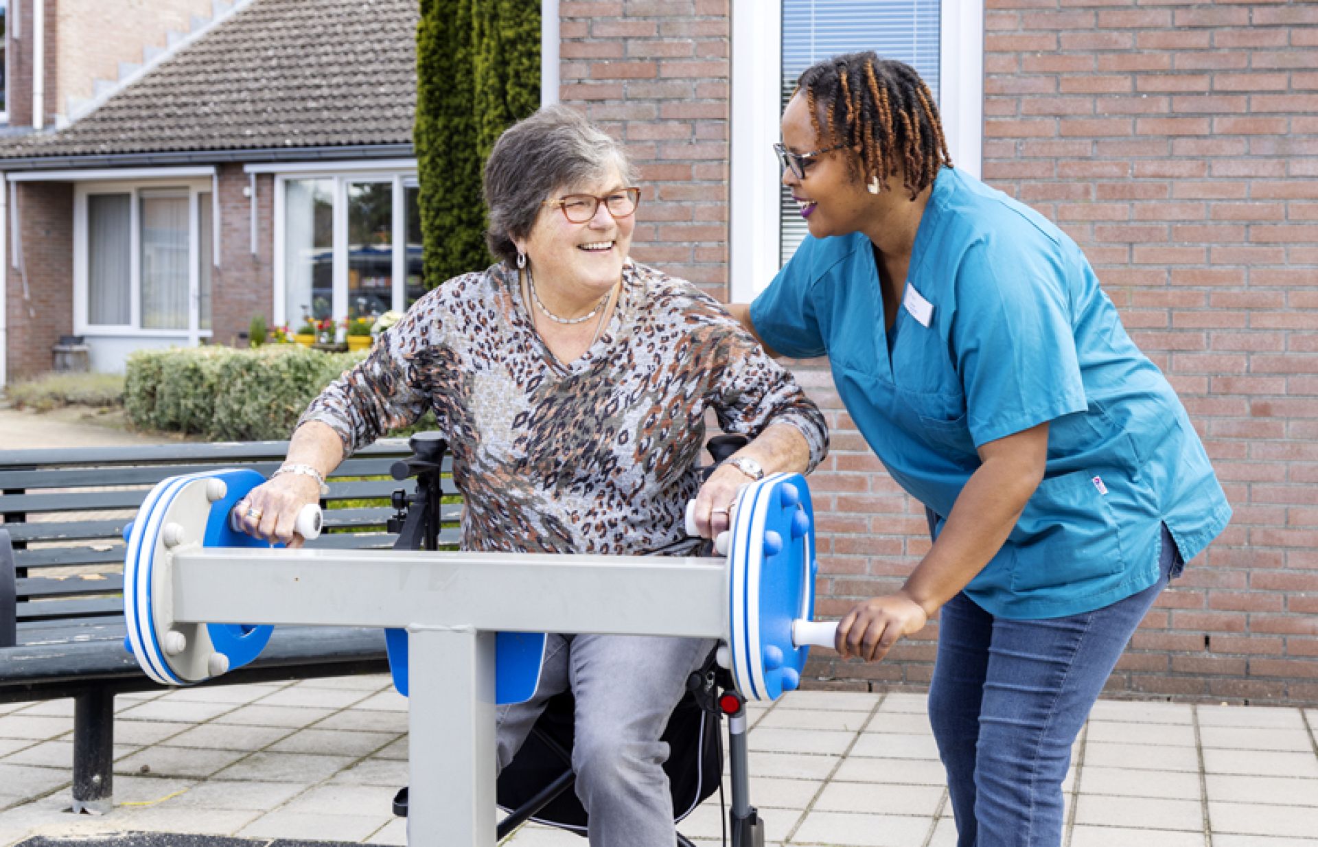 Een bewoner is buiten aan het sporten op een beweegapparaat en krijgt daarbij hulp van een zorgmedewerker