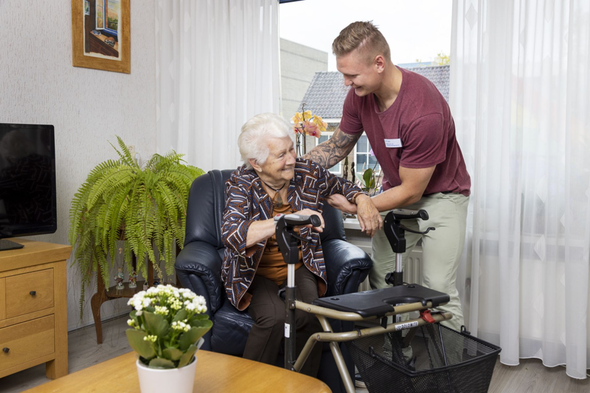 Bewoner krijgt hulp van een medewerker bij het opstaan uit haar stoel.