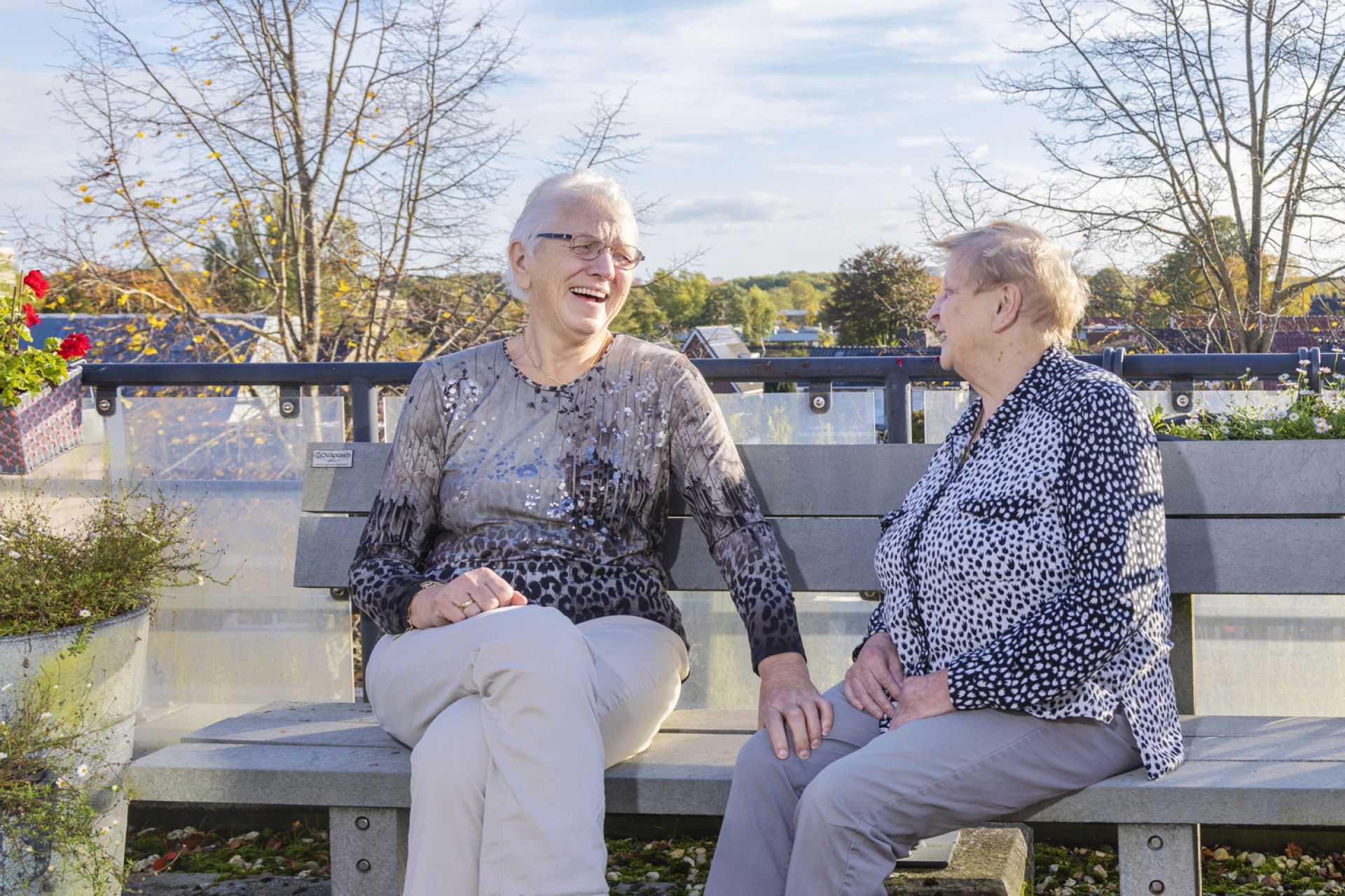 Twee bewoners zitten lachend buiten op een bankje in de zon
