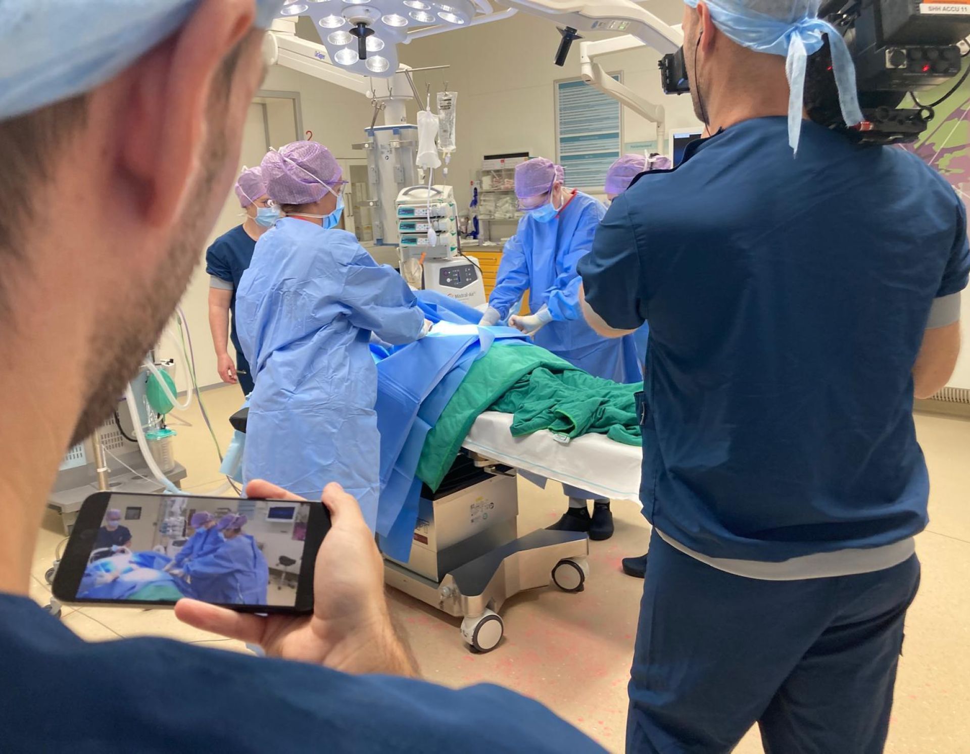 Medisch team voert een operatie uit in een operatiekamer, terwijl een persoon op de voorgrond een opname maakt met een smartphone