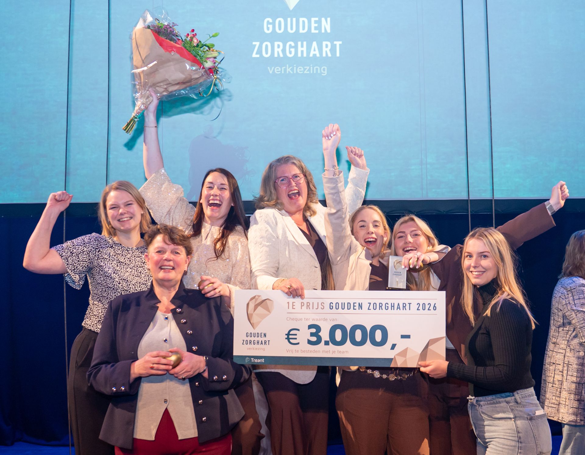 Collega's van woonzorgcentrum Holdert in Emmen juichen enthousiat met de cheque van 3000 euro in de hand en een bos bloemen