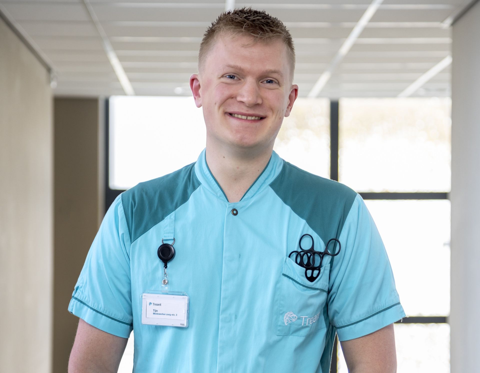 Een man in een lichtblauw uniform staat glimlachend in een gang.